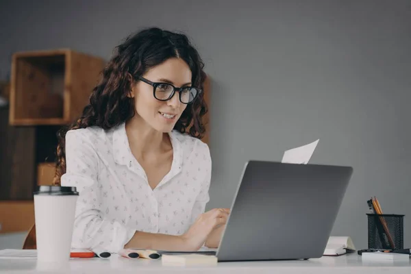 Jovem mulher europeia feliz vestindo óculos em camisa branca trabalhando no laptop moderno de casa — Fotografia de Stock