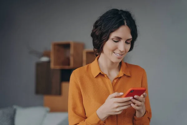 Молодая улыбающаяся латиноамериканка держит смартфон, смотрит на экран с улыбкой и смс — стоковое фото