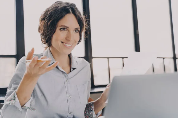 Mujer alegre tutor español mira a la pantalla del ordenador portátil con sonrisa segura al explicar un nuevo tema —  Fotos de Stock