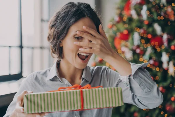 Νέοι ενθουσιασμένοι ισπανική γυναίκα κοιτάζοντας μέσα από τα δάχτυλα στο δώρο Χριστούγεννα, ενώ γιορτάζει τα Χριστούγεννα στο σπίτι — Φωτογραφία Αρχείου
