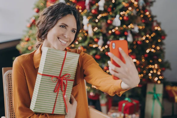 Χαρούμενη Ιταλίδα που κάθεται κοντά στο όμορφο χριστουγεννιάτικο δέντρο στο σπίτι και βγάζει selfie με δώρο — Φωτογραφία Αρχείου
