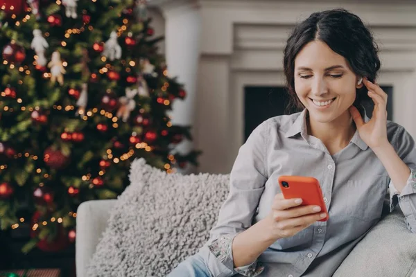 リラックスした幸せなイタリア人女性座っています居心地の良いソファに自宅で電話でクリスマス休暇中 — ストック写真