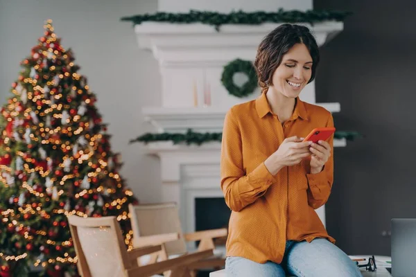 Arka planda Noel ağacı olan bir evde otururken cep telefonu kullanan neşeli Avrupalı kadın