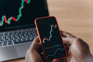 Afrikalı bir adamın erkek elleri akıllı telefonu bilgisayarın önünde tutuyor ve üzerinde ticaret çizelgeleri var.