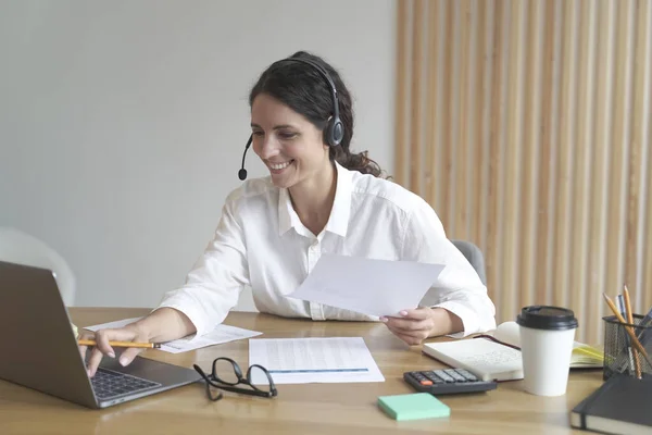 ハッピー女性は自宅のオフィスで机の上に座っている間にヘッドフォンを身に着けているラップトップ画面で動作しますオンライン — ストック写真