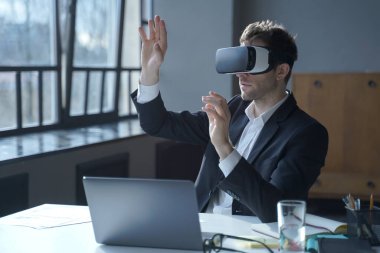 VR kulaklıklı erkek ofis çalışanı masada otururken dijital arayüzle etkileşime giriyor