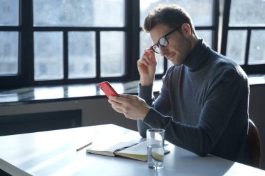 Çekici Avrupalı erkek girişimci elinde cep telefonuyla ofis masasında oturuyor