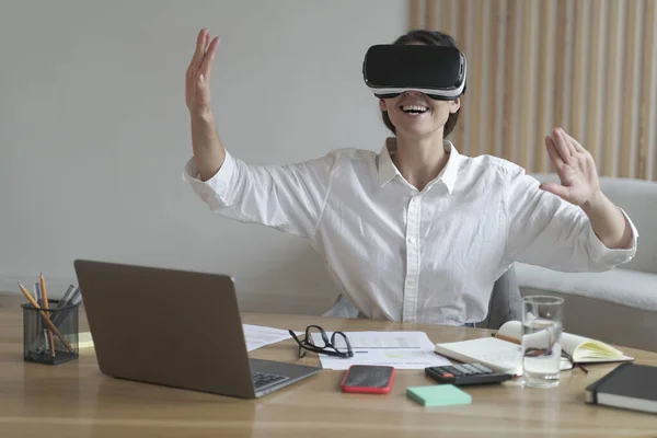 Joven mujer de negocios alegre moviendo las manos en el aire mientras usa gafas de realidad virtual en el lugar de trabajo — Foto de Stock