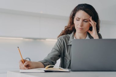 Üzgün, yorgun, serbest çalışan biri evde çalışıyor. Yorgun Avrupalı kadın dizüstü bilgisayarın önünde oturuyor ve not alıyor. Çok çalışan kızın başı ağrıyor. Uzaktan çalışma stresi kavramı.