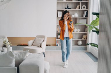 Kulaklıklı mutlu Avrupalı kızın evde telefonu var. Genç kadın kapalı alanda hava podu kullanıyor. Kablosuz kulaklıklar. Rahatlama, rahatlama ve iletişim konsepti. Güzel modern iç mekan.