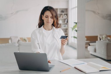 Beyazlı genç güzel kadın internetten satın alacak. Avrupalı bir kız dizüstü bilgisayarın önünde kredi kartı tutuyor. İnternetten rezervasyon ya da satın alma. E-ticaret ve mali işlem kavramı.
