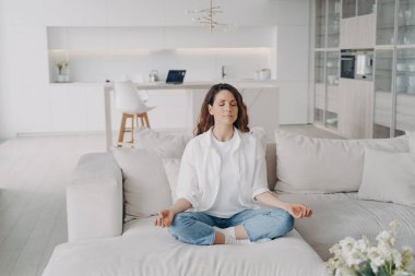 Genç İspanyol kadın evdeki kanepede yoga ve meditasyon yapıyor. Duruş egzersizi, Lotus Asana. Sabah jimnastiği, sağlıklı yaşam tarzı. Konsantrasyon ve zen bulma kavramı.