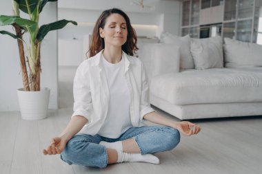 Kafkasyalı genç bir kadın Lotus Asana 'da oturuyor. Kız evde yoga ve meditasyon yapıyor. Sabah jimnastiği. Uyum ve zihin rahatlatma kavramı. Oturma odasının İskandinav iç mekanı.