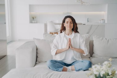 Huzurlu genç Latin kadın evdeki kanepede yoga ve meditasyon yapıyor. Çekici kız Lotus Asana 'da duruş egzersizi yapıyor. Oturma odasında sabah jimnastiği. Denge, zen ve uyum.