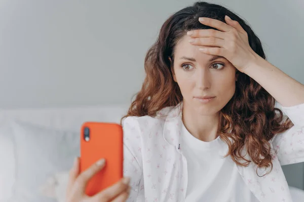 Worried girl is reading trouble news looking at smartphone screen. Young attractive woman is chatting on phone or browsing internet at home. European girl is getting stress and anxiety attack.