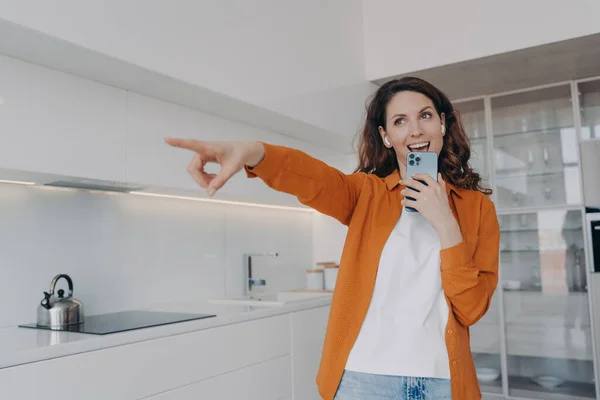 Carefree spanish woman in earphones is singing with cellphone as with microphone. Application for music listening online. Playful passionate girl is singing at home and points to you.