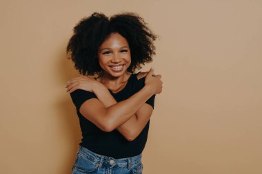 Güzel, mutlu, koyu tenli, şehvetli afro saç stili, kendini ışıltılı bir gülümseme ile kucaklayan, stüdyo bej arka planında fotokopi alanı ile izole edilmiş bir kadın görüntüsü. Kişisel aşk kavramı