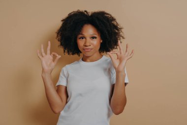 Her şey yolunda. Beyaz tişörtlü Afro-Amerikalı genç bayan iki elini de iyi bir jestte tutuyor. Stüdyoda kahverengi arka planda tek başına dururken 