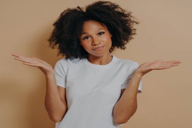 Tereddütlü bir şekilde avuç içi kaldıran, kafası karışmış kameraya bakan, melez bir kadın, emin olmayan ve bej duvara karşı poz verirken bazı şüpheleri olan genç Afrikalı bir kadın.