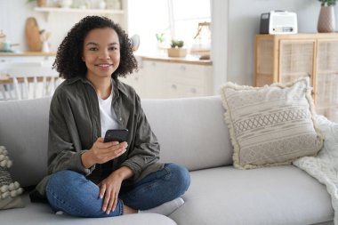 Young hispanic woman is browsing social media and smiling. Curly african american girl has fun with phone at home. Chatting with friends remotely. Lazy weekend morning. Online book reading.
