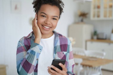 Modern mutlu, çift ırklı, kablosuz kulaklıklı genç kız elinde akıllı telefon, müzik dinliyor, radyo şarkısı dinliyor. Gülümseyen melez kadın melodi sesinden zevk alıyor, evde müzik uygulamaları kullanıyor..