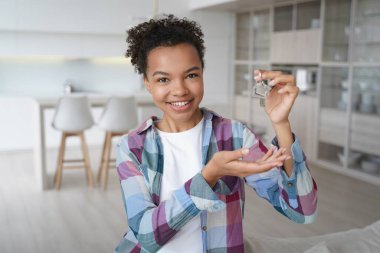 New apartment buyer. Happy teenager is homeowner in living room of new residence. Carefree african american girl is holding house key and smiling. Mortgage loan and relocation conceptual image.