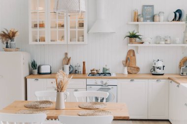 İskandinav mutfak tasarımı. Modern apartman dekorasyonu. Ev mutfağı ya da yemek odası. Beyaz, lüks iç mekan gün ışığında aydınlanıyor. Mutfak aletleri, mutfak gereçleri, soba, masa ve buzdolabı..