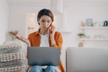 Young caucasian woman has laptop problem. Girl has consultation on phone. Lady is talking on telephone and telling some problem. Puzzled woman is sitting on sofa at home holding computer.