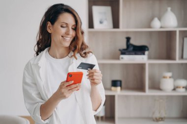 Akıllı telefon ve kartla kolay ödeme. Mutlu Kafkasyalı kız evde internetten satın alacak. Kız internetten yer ayırtıyor ya da alıyor. E-ticaret ve mali işlem kavramı.