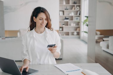 Happy spanish girl is holding credit card in front of pc. Young pretty woman is going to buy through internet. Girl is booking or purchasing online. E-commerce and financial transaction concept.