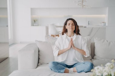Serene latin woman at home. Posture exercise, lotus asana. Young european woman is practicing yoga and meditation on couch with her legs crossed. Minimal white modern scandinavian interior.