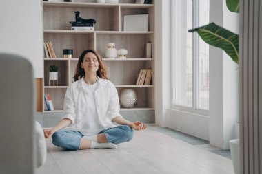 Sakin, mutlu Latin kadın evde yoga ve meditasyon yapıyor. Kız sabah Lotus Asana 'da pencerenin yanında oturuyor. Sabah jimnastiği. Stres atma ve zihin dengesi kavramı.