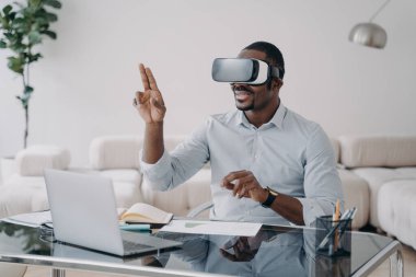 VR gözlüklü heyecanlı afro adam iş yerinde. İş adamının sanal toplantısı var. Freelancer tasarım projesi üzerinde çalışıyor. Uzak çalışma ve eğlence için modern kablosuz teknoloji.