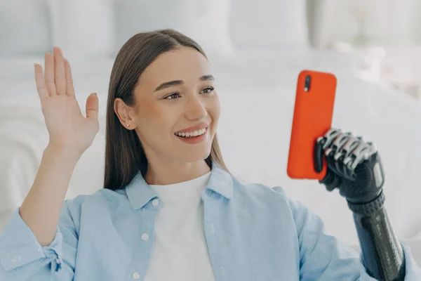 Young disabled woman has video call on phone. European girl is holding the mobile phone with bionic artificial arm and taking selfie. Happy caucasian woman with artificial limb at home.