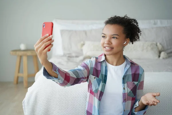 Morning video phone call. Young african american woman is talking to friend on smartphone in bedroom. Teenage girl is holding smartphone and speaking in front of camera. Phone addiction concept.