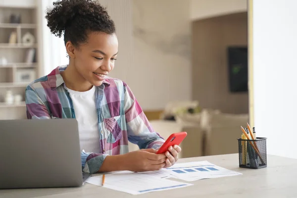 Afro teenage girl is distracting while studying remote. Teenage girl sitting in front of laptop, clicks mobile phone and chatting. African american student gets remote education at school or college.