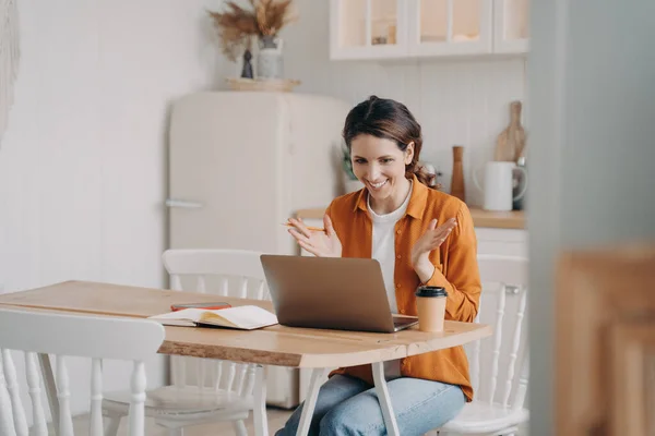 Friendly lady has business video call by computer at home. Caucasian young woman in orange shirt is remote worker sitting at table at kitchen. Successful entrepreneur. Freelance distance work concept.