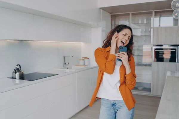 Playful girl is dancing alone at home. Excited european woman in airpods is singing and screaming with mobile telephone as with mic. App for music listening online. Happy housewife having fun.