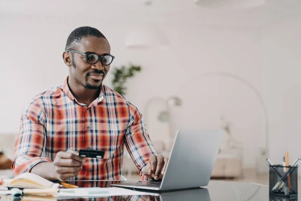 Happy african american man in glasses is purchasing online from home using laptop and credit card. Internet banking and money transfer. Ecommerce and consumerism concept.