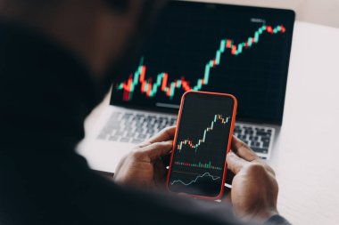 Borsa konsepti. Akıllı telefon tutan kendinden emin Afro-Amerikan işadamının kesilmiş fotoğrafı, finansal istatistiklerle çalışıyor, telefon ve dizüstü bilgisayarlardaki şemaları karşılaştırıyor, veri analizi yapıyor.