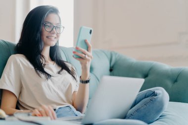 Olumlu iyimser esmer kadın günlük kıyafetler ve optik gözlükler giymiş, akıllı telefon ekranına odaklanmış, yeni bir web sitesi arıyor, internetten eğitim Webinar 'ını izliyor, kanepeye oturuyor.