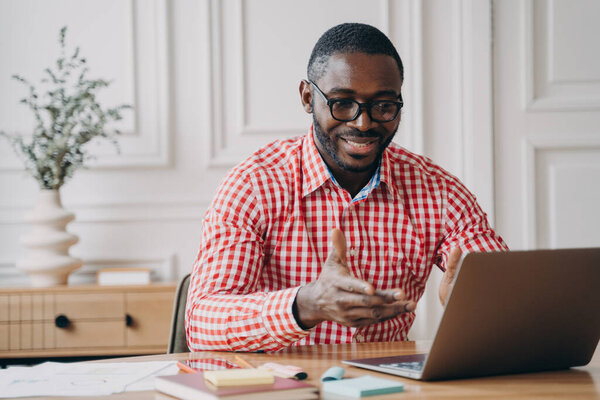 Positive african man online teacher tutor looking at laptop screen and smiling, talking with students while working remotely from home, dressed in casual clothes. E-learning and distant education