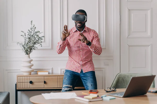 Young programmer or software developer African American man in VR headset glasses testing 3D games and applications standing in modern home office interior. Technology and software development concept