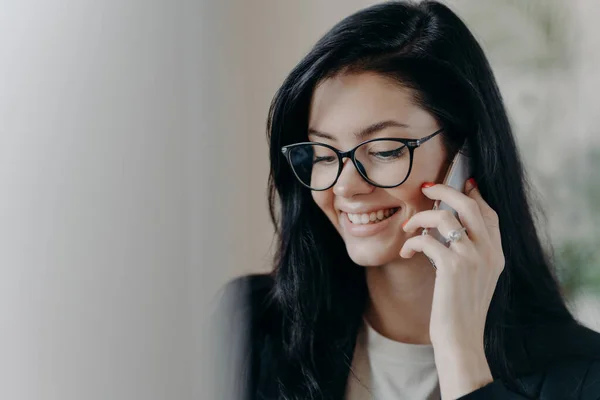 Happy woman lawyer keeps mobile phone near ear, has pleasant conversation with client, discuss working issues, smiles gently, wears transparent glasses, formal wear. Female employee calls someone