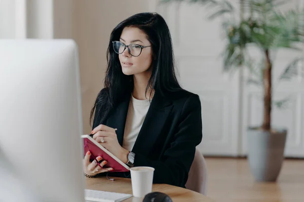 Indoor shot of prosperous businesswoman makes notes in notepad, concentrated in computer screen, watches training video how to start own business, poses at desktop with coffee cup, makes report