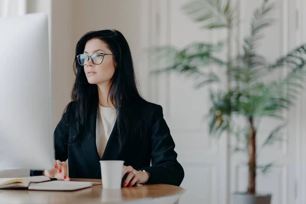 Concentrated female entrepreneur wears elegant black costume, works at computer, sits at table in cozy work place wears transparent glasses browses internet makes business project thinks over strategy