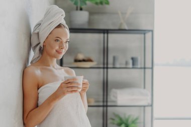 Banyo havlusuna sarılı, bir kupa çayla poz veren, uzaklığa odaklanmış, iç duvarın yanında duran, sıcacık atmosferden hoşlanan güzel genç bir kadının yatay görüntüsü.