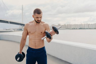 Güçlü fitness sportif kıllı sakallı adam dambıllarıyla egzersiz yapıyor, pazılarını şişiriyor, dışarıda poz veriyor, çıplak gövdesi var, temiz hava soluyor. Fitness, spor ve yaşam tarzı konsepti.