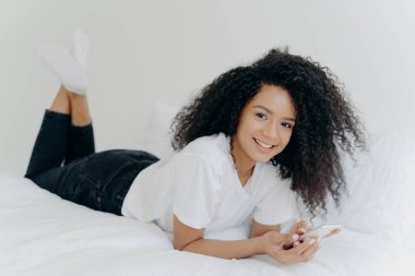 Bin yıllık neşeli Afro saçlı bir kadının kapalı çekimleri, rahat bir yatakta karnı üzerine uzanmış, sohbet ediyor, e-posta kutusunu kontrol ediyor, beyaz tişört, çorap ve siyah pantolon giyiyor, tembel bir öğleden sonra geçiriyor.
