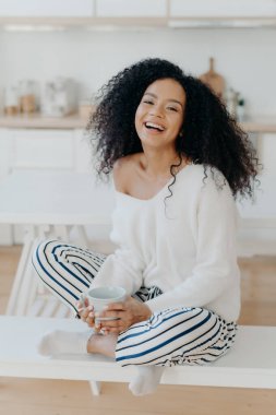 Vertical shot of pleased young Afro American woman feels relaxed and carefree, drinks aromatic coffee, sits crossed legs against kitchen interior, being in good mood, spends spare time at home